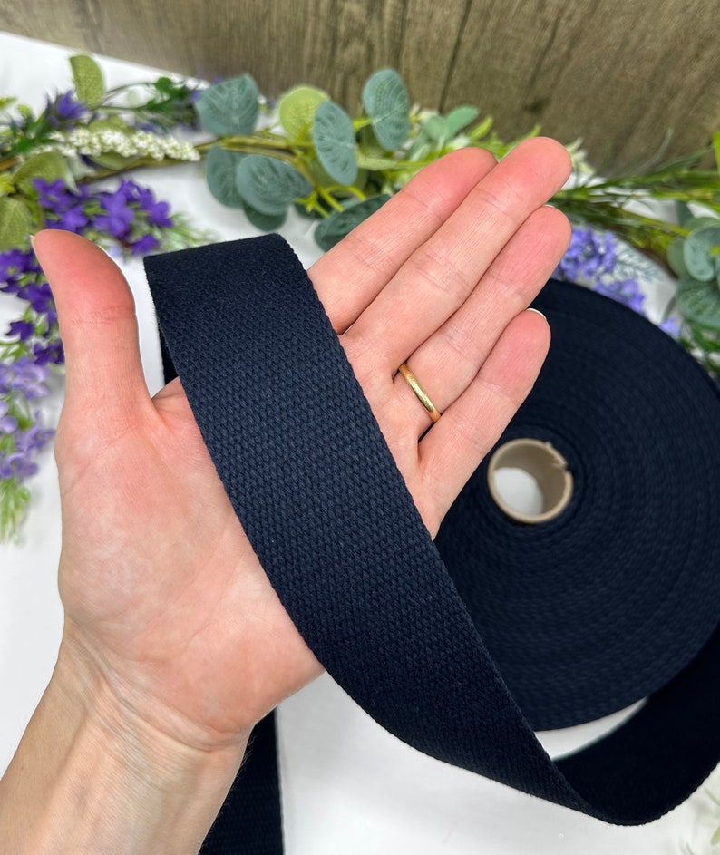 May include: A roll of black cotton webbing, approximately 1 inch wide, is shown in the image. The webbing is a woven fabric with a smooth texture. The webbing is being held in a person's hand.