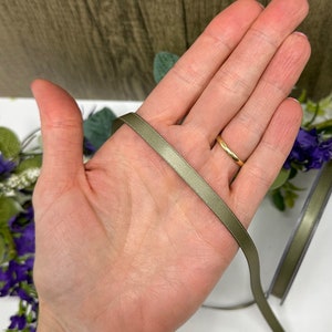 May include: A close-up of a hand holding a sage green satin ribbon. The ribbon is about 1/2 inch wide and has a smooth, silky texture.