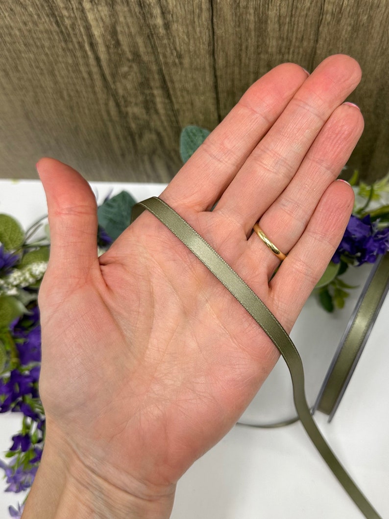 May include: A close-up of a hand holding a sage green satin ribbon. The ribbon is about 1/2 inch wide and has a smooth, silky texture.