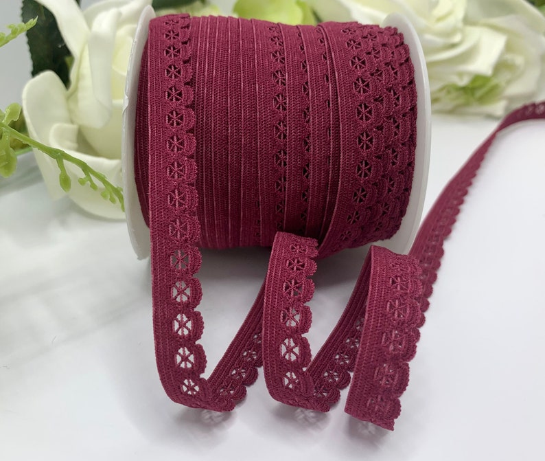 Puede incluir: Un rollo de encaje el&aacute;stico de color burdeos con borde festoneado. El encaje tiene un delicado patr&oacute;n floral.