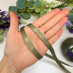 May include: A close-up of a hand holding a length of olive green satin ribbon. The ribbon is folded in half and the ends are visible.