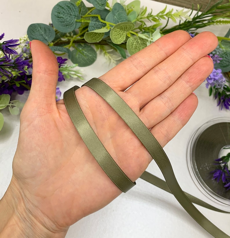 May include: A close-up of a hand holding a length of olive green satin ribbon. The ribbon is folded in half and the ends are visible.