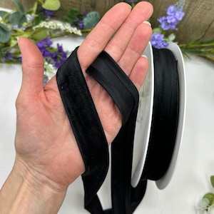 K&ouml;nnte beinhalten: Schwarzes Samtband mit weicher, glatter Textur. Das Band ist in der Mitte gefaltet und wird in einer Hand gehalten. Das Band ist etwa 2,5 cm breit.
