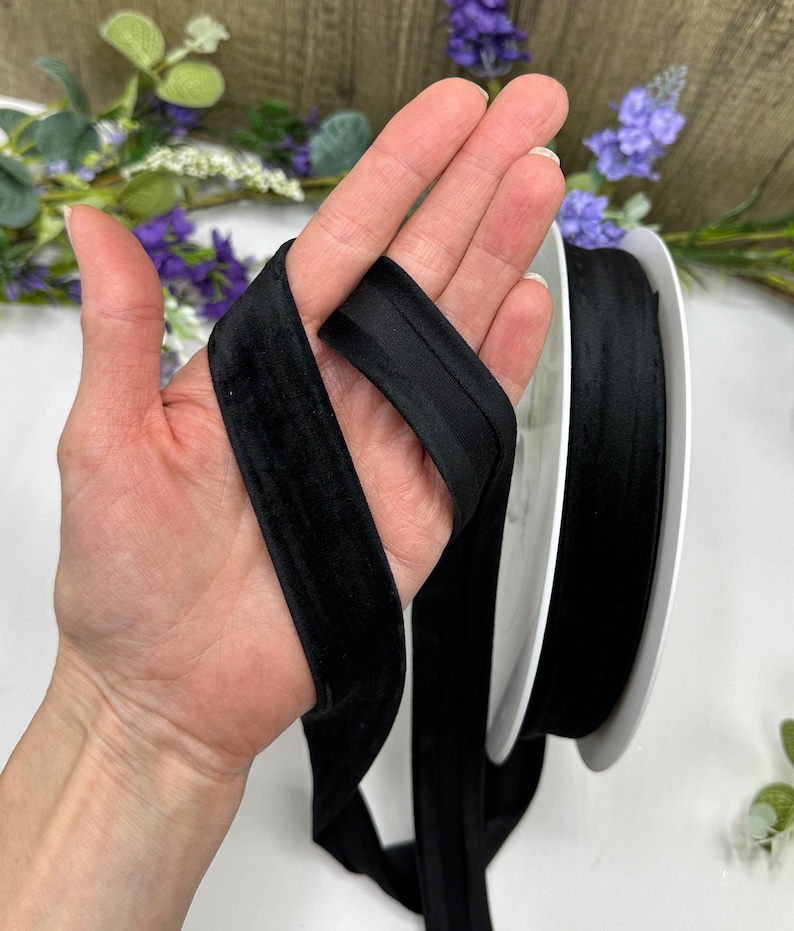 K&ouml;nnte beinhalten: Schwarzes Samtband mit weicher, glatter Textur. Das Band ist in der Mitte gefaltet und wird in einer Hand gehalten. Das Band ist etwa 2,5 cm breit.
