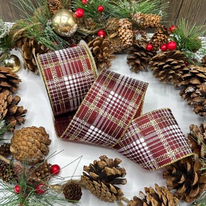 Puede incluir: Un rollo de cinta a cuadros burdeos y blanca con ribete dorado, desenrollada para mostrar el patrón. La cinta está rodeada de piñas, vegetación y bayas rojas, creando un tema festivo navideño.