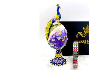 May include: A purple and gold peacock figurine with a jeweled crown on its head. The peacock is perched on a purple and gold egg-shaped base. The figurine is next to a black box with gold lettering that reads "Erre Lorre Jewellers".