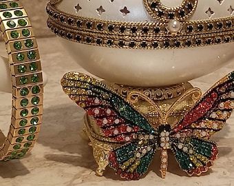 May include: A white egg-shaped music box with a gold and black trim and a jeweled butterfly on the base. The butterfly has red, green, yellow, and blue jewels.