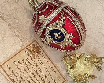 May include: A red and gold Faberge egg replica with a blue and gold crest. The egg is on a gold stand and has a card that reads "Pierre Lorren Authenticity Card Luxurious 5 star Russian Faberge egg. Classy Handmade Trinkets by Master Jewellers of Cyprus 24k Gold decor - Swarovski Diamonds Balance the Imperial Lion Egg to ensure Luck, Happiness, Health, Love, Strength & Courage comes your way!"