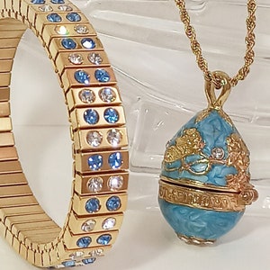 Puede incluir: Un collar de cadena dorada con un colgante en forma de huevo azul y dorado. El colgante tiene un dise&ntilde;o floral y est&aacute; decorado con peque&ntilde;os cristales azules y blancos. El collar se combina con una pulsera dorada con cristales azules y blancos.