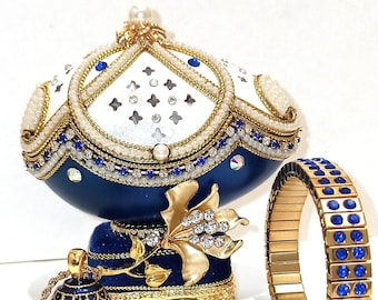 May include: A blue and white jeweled egg-shaped music box with a gold base and a matching blue and gold pendant necklace and bracelet. The music box is decorated with pearls and rhinestones.