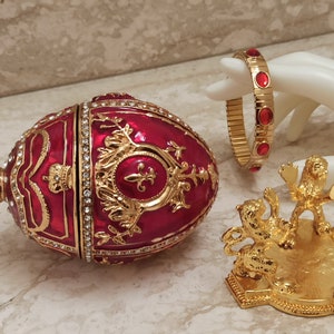 Op de afbeelding: Een rood en gouden juwelen eivormig doosje met een kroon erop. Het doosje staat op een gouden standaard met drie leeuwenfiguren.