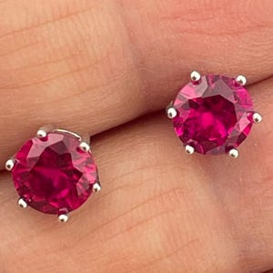 Könnte beinhalten: Ein Paar Ohrstecker mit großen, runden, leuchtend magentafarbenen Edelsteinen, die in silberfarbenem Metall gefasst sind. Die Ohrringe werden vor einem roten Hintergrund gezeigt.