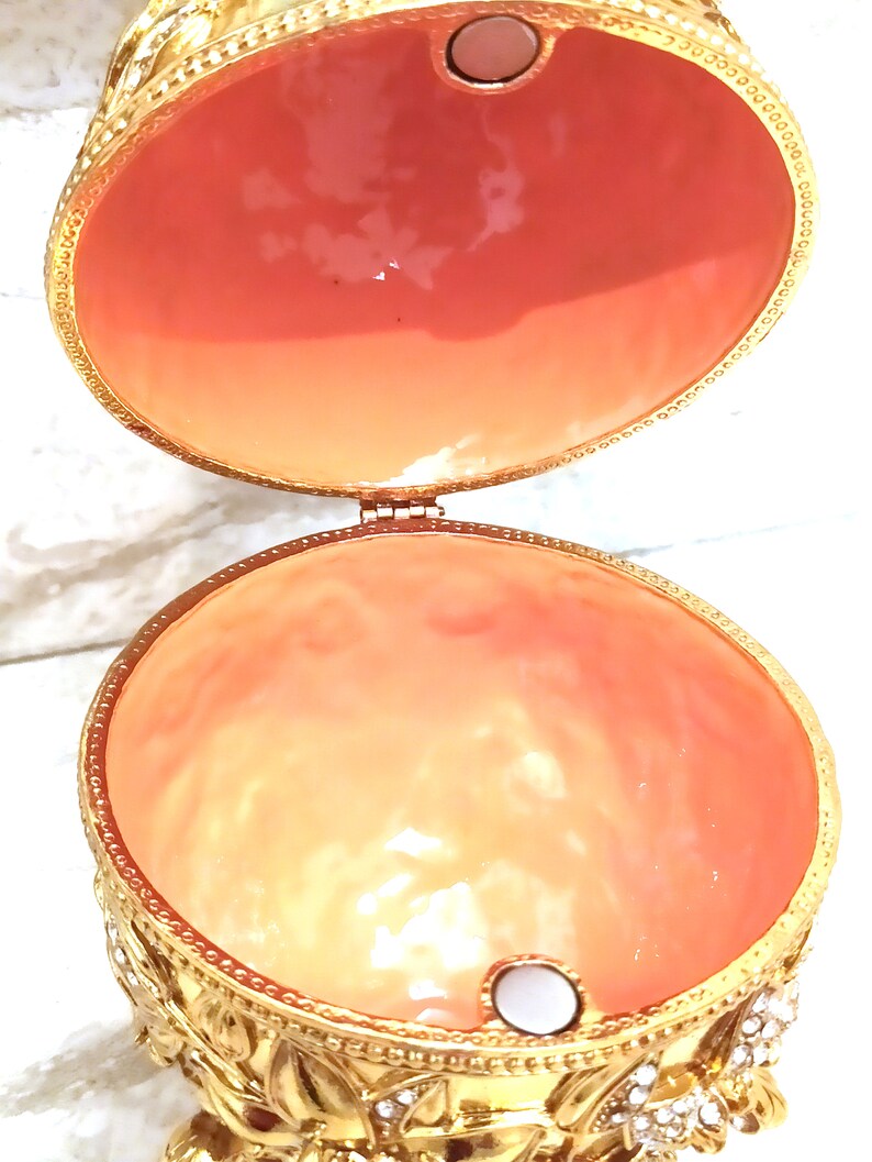 Puede incluir: Una caja de alhajas en forma de huevo de color dorado con una tapa con bisagras. El interior de la caja est&aacute; forrado con un material de color melocot&oacute;n. La caja est&aacute; decorada con detalles intrincados y tiene un peque&ntilde;o bot&oacute;n blanco nacarado en la parte inferior.