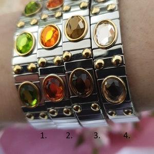 May include: Four stackable bracelets with different coloured gemstones. The bracelets are made of silver metal with gold accents. The gemstones are green, orange, brown, and clear.