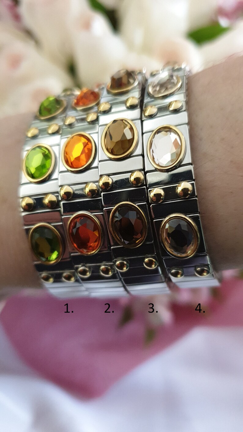 May include: Four stackable bracelets with different coloured gemstones. The bracelets are made of silver metal with gold accents. The gemstones are green, orange, brown, and clear.