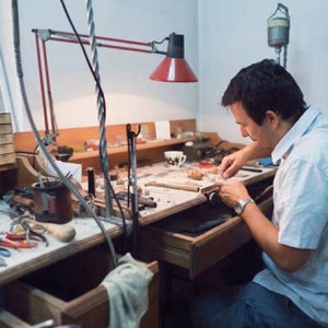 May include: A person working at a workbench with a variety of tools and materials. The workbench is made of wood and has a metal frame. The person is wearing a white shirt and is using a small tool to work on a piece of jewellery.