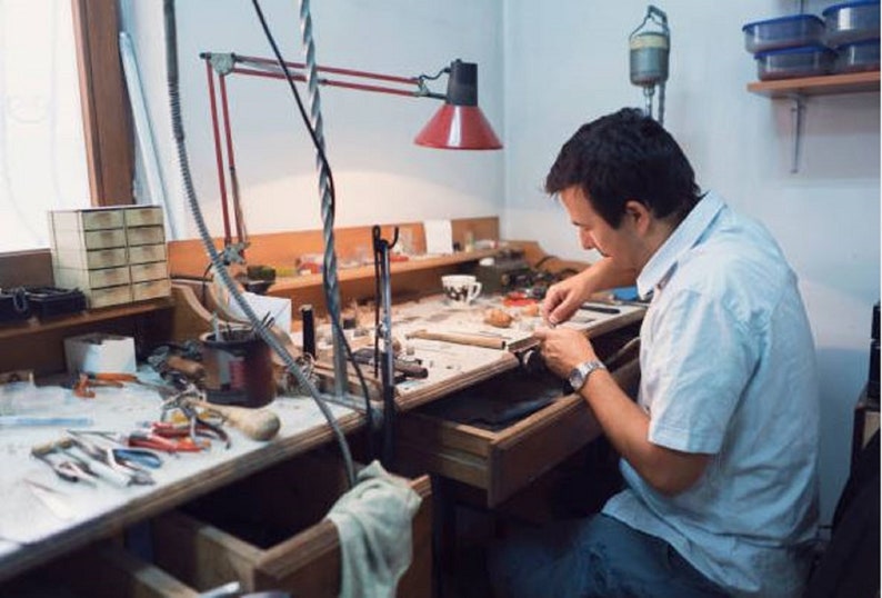 May include: A person working at a workbench with a variety of tools and materials. The workbench is made of wood and has a metal frame. The person is wearing a white shirt and is using a small tool to work on a piece of jewellery.