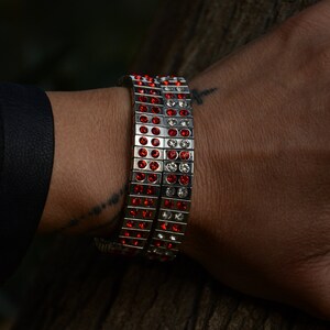 May include: A silver bracelet with red and clear crystals. The bracelet is made of multiple rows of small squares, each with a crystal in the centre.