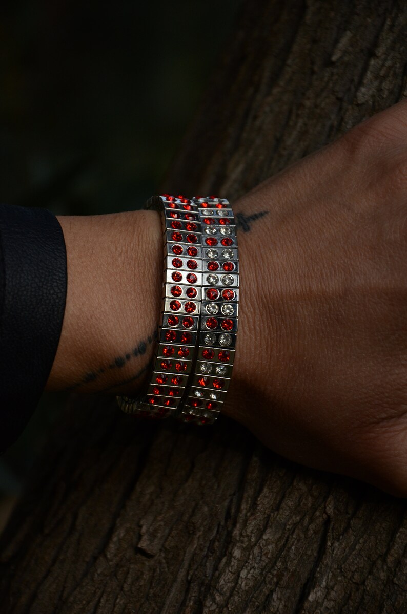 May include: A silver bracelet with red and clear crystals. The bracelet is made of multiple rows of small squares, each with a crystal in the centre.