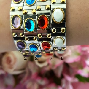 May include: A stack of four bracelets featuring different coloured gemstones. The bracelets are made of silver metal with gold accents. The gemstones are purple, blue, red, and white.