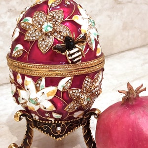 Puede incluir: Un huevo rojo y dorado adornado con joyas con un dise&ntilde;o de abeja y un patr&oacute;n floral. El huevo est&aacute; en un soporte y al lado de una granada roja.