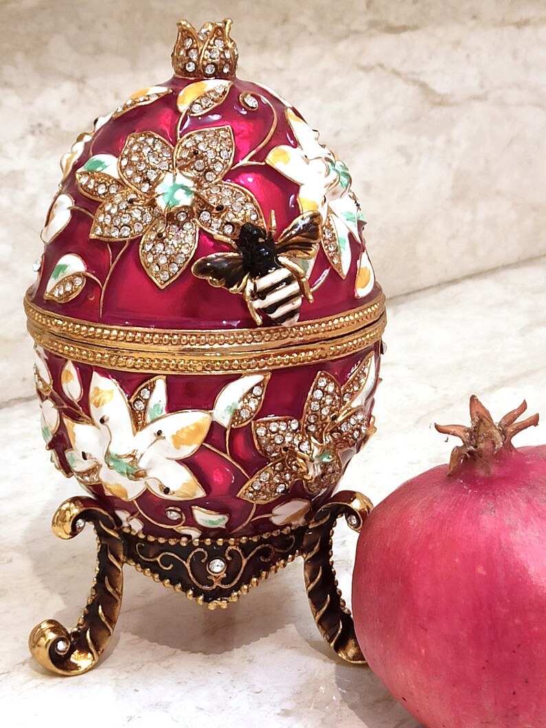 Puede incluir: Un huevo rojo y dorado adornado con joyas con un dise&ntilde;o de abeja y un patr&oacute;n floral. El huevo est&aacute; en un soporte y al lado de una granada roja.