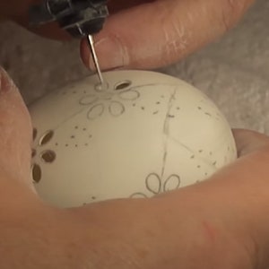 May include: A person is using a small tool to carve a design into a white egg. The design includes a series of small flowers and dots.