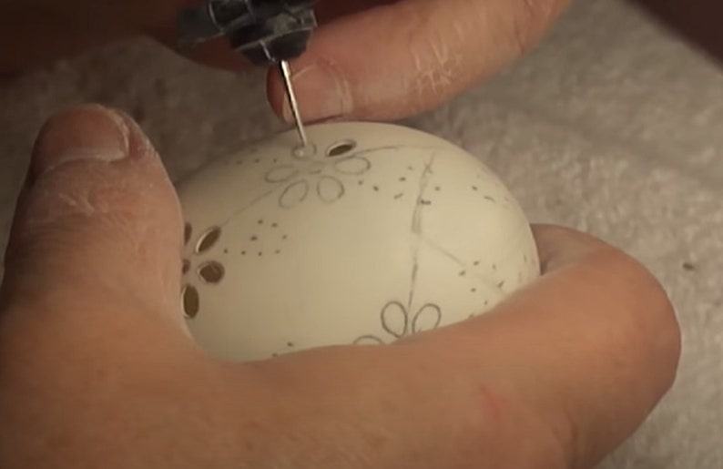 May include: A person is using a small tool to carve a design into a white egg. The design includes a series of small flowers and dots.
