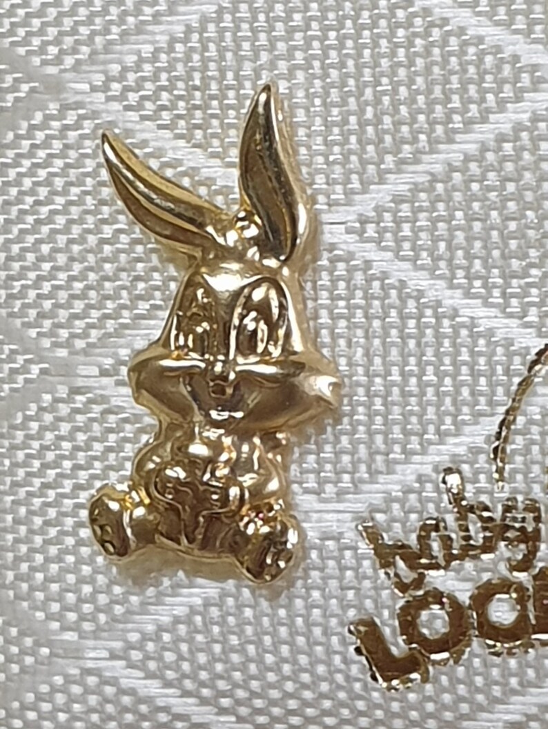 May include: A gold-colored Bugs Bunny charm. The charm is three-dimensional and shows Bugs Bunny sitting down with his hands in his lap.