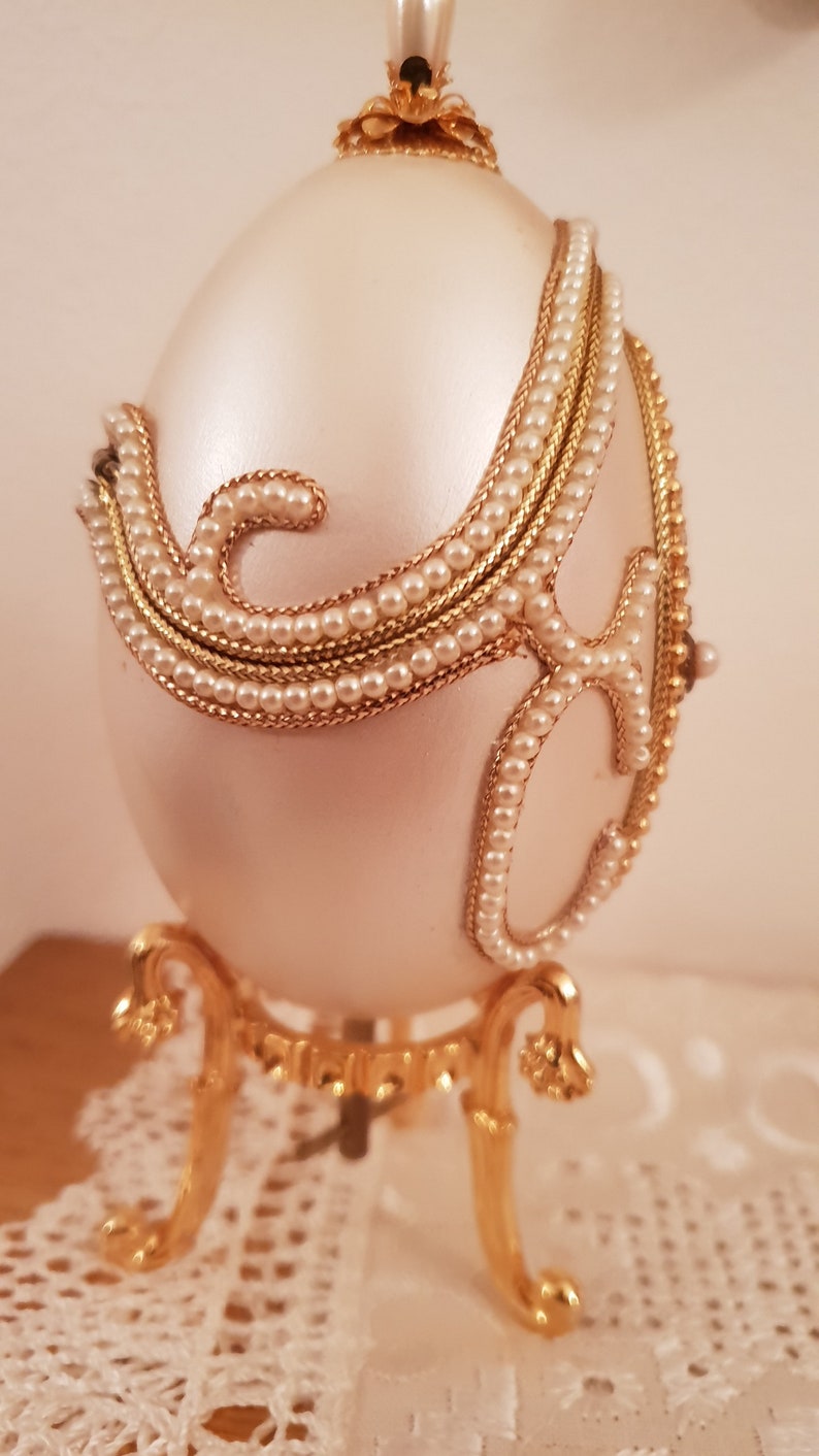 May include: A white porcelain egg with gold and pearl accents. The egg is on a gold stand with three legs.