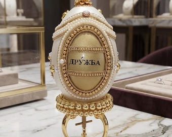 May include: An ornate, egg-shaped decorative object in cream and gold, featuring a central oval panel with the Cyrillic text "ДРУЖБА". The egg is adorned with rows of small white pearls, gold filigree, and a pearl finial, resting on a gold stand.