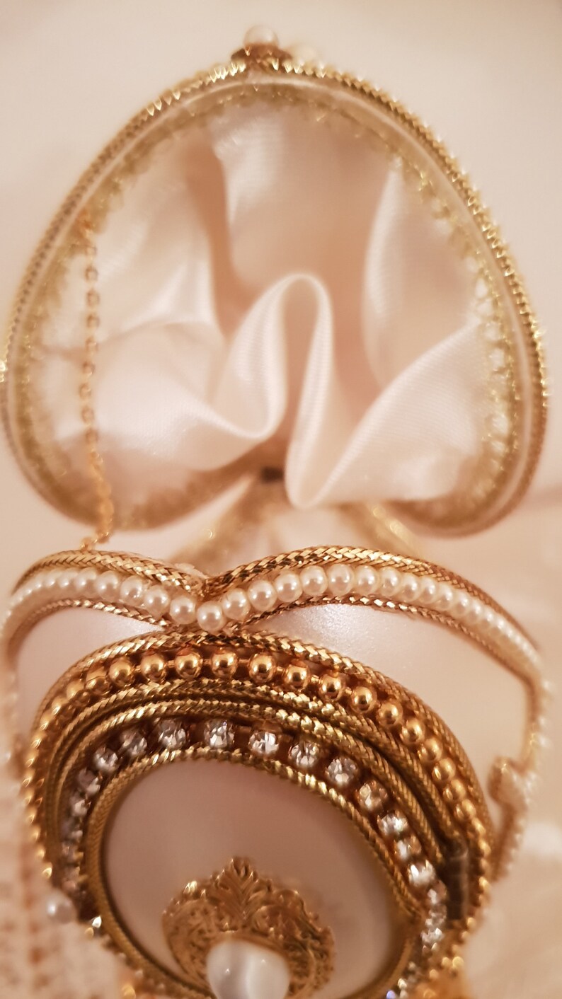 May include: A white egg-shaped trinket box with a gold and pearl trim. The box is open and the inside is lined with white satin.