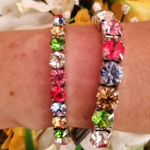 May include: Two silver bracelets with colourful crystal stones. The bracelets are on a person's wrist. The stones are arranged in a rainbow pattern.