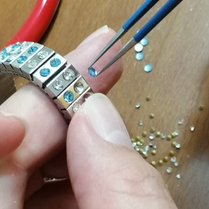 May include: A silver bracelet with blue and clear rhinestones is being assembled with tweezers. A small blue rhinestone is being placed on the bracelet.
