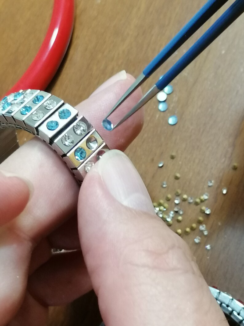 May include: A silver bracelet with blue and clear rhinestones is being assembled with tweezers. A small blue rhinestone is being placed on the bracelet.
