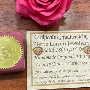 May include: A gold certificate of authenticity for a Looney Tunes Warner Bros. 18 karat gold pendant. The certificate is in a red velvet box with a gold seal. The text on the certificate reads: "Certificate of Authenticity Pierre Lorren Jewellery Solid 18kt GOLD Handmade Original Vintage Looney Tunes Warner Bros 2000 edition by Master Jewellers in Cyprus For Centuries Gold has symbolised Royalty, Health & Prosperity. Truly a luxury gift for someone special!"