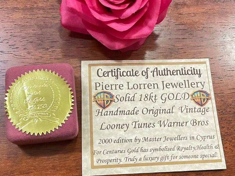 May include: A gold certificate of authenticity for a Looney Tunes Warner Bros. 18 karat gold pendant. The certificate is in a red velvet box with a gold seal. The text on the certificate reads: "Certificate of Authenticity Pierre Lorren Jewellery Solid 18kt GOLD Handmade Original Vintage Looney Tunes Warner Bros 2000 edition by Master Jewellers in Cyprus For Centuries Gold has symbolised Royalty, Health & Prosperity. Truly a luxury gift for someone special!"