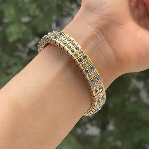 Puede incluir: Un brazalete dorado con cristales azules y transparentes. El brazalete est&aacute; hecho de peque&ntilde;os cuadrados unidos.