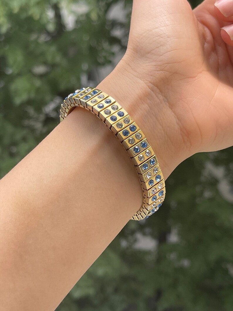 Puede incluir: Un brazalete dorado con cristales azules y transparentes. El brazalete est&aacute; hecho de peque&ntilde;os cuadrados unidos.