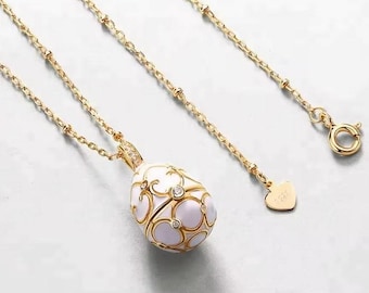 Pode incluir: Um colar de corrente de ouro com um pingente de ovo de esmalte branco. O ovo é decorado com filigrana dourada e pequenas pedras transparentes.