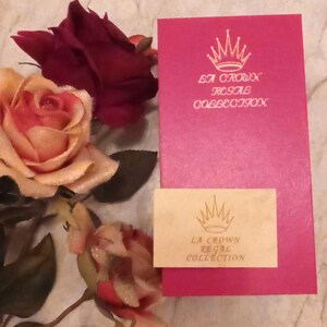 Puede incluir: Una caja rosa con una corona dorada y el texto "La Crown Regal Collection" en la parte delantera. La caja est&aacute; sobre una superficie blanca con una rosa.