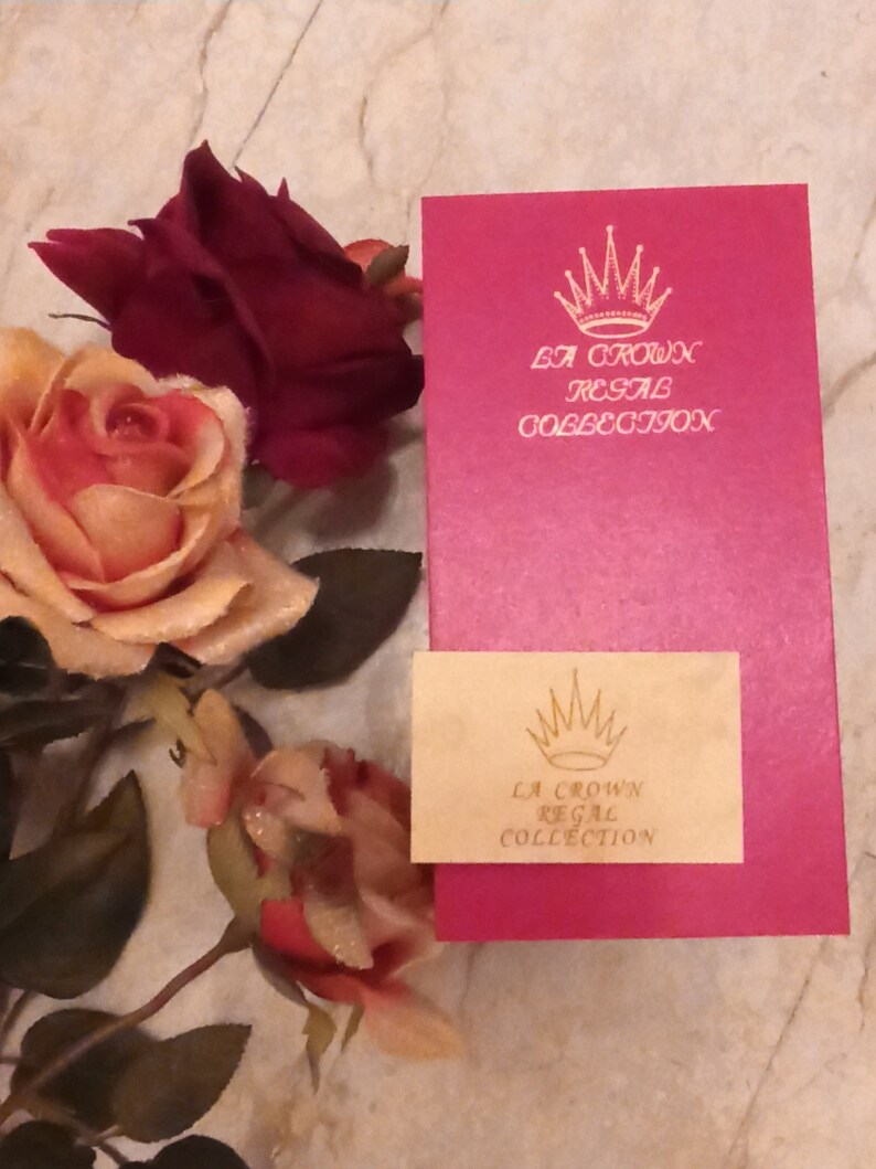 Puede incluir: Una caja rosa con una corona dorada y el texto "La Crown Regal Collection" en la parte delantera. La caja est&aacute; sobre una superficie blanca con una rosa.