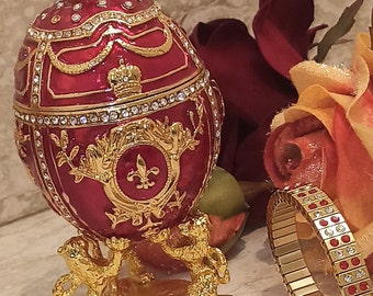 May include: A red and gold jeweled egg with a crown on top, sitting on a gold stand with two golden animal figures. The egg is decorated with a fleur-de-lis and other intricate details.