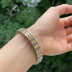 Puede incluir: Un brazalete dorado con una doble fila de cristales azules y transparentes. El brazalete est&aacute; hecho de peque&ntilde;os cuadrados unidos.