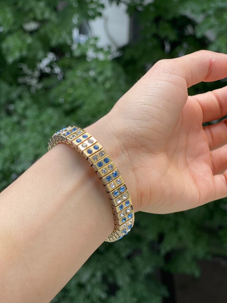 Puede incluir: Un brazalete dorado con una doble fila de cristales azules y transparentes. El brazalete est&aacute; hecho de peque&ntilde;os cuadrados unidos.