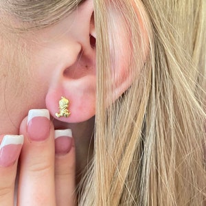 May include: Gold-toned stud earrings featuring a small cartoon character design. The earrings are worn in a person's earlobe.