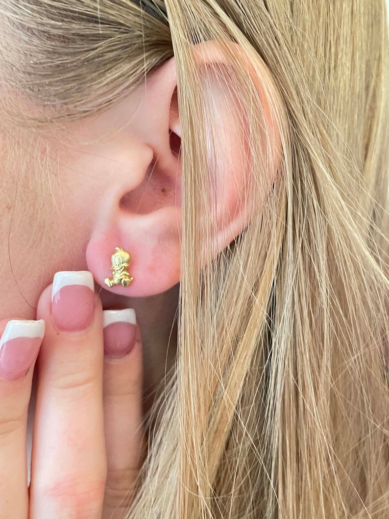 May include: Gold-toned stud earrings featuring a small cartoon character design. The earrings are worn in a person's earlobe.