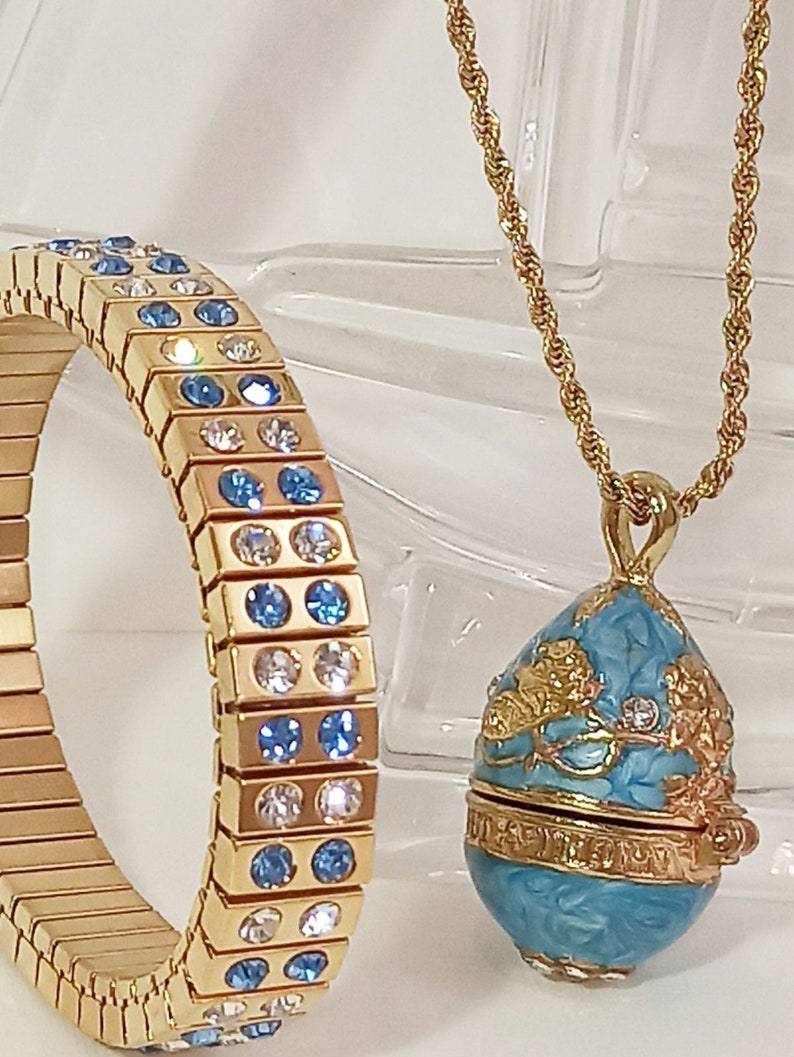 Puede incluir: Un collar de cadena dorada con un colgante en forma de huevo azul y dorado. El colgante tiene un dise&ntilde;o floral y est&aacute; decorado con peque&ntilde;os cristales azules y blancos. El collar se combina con una pulsera dorada con cristales azules y blancos.