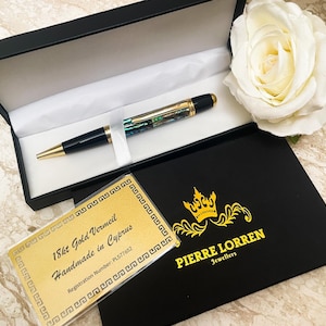 May include: A luxury pen in a black presentation box. The pen features a gold-colored nib and accents, with a black barrel and a decorative iridescent section. A white rose and a black box with gold text are also in the image.