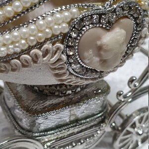May include: A white porcelain egg decorated with pearls and rhinestones, set in a silver carriage with wheels. The egg has a heart-shaped opening with a cameo inside.